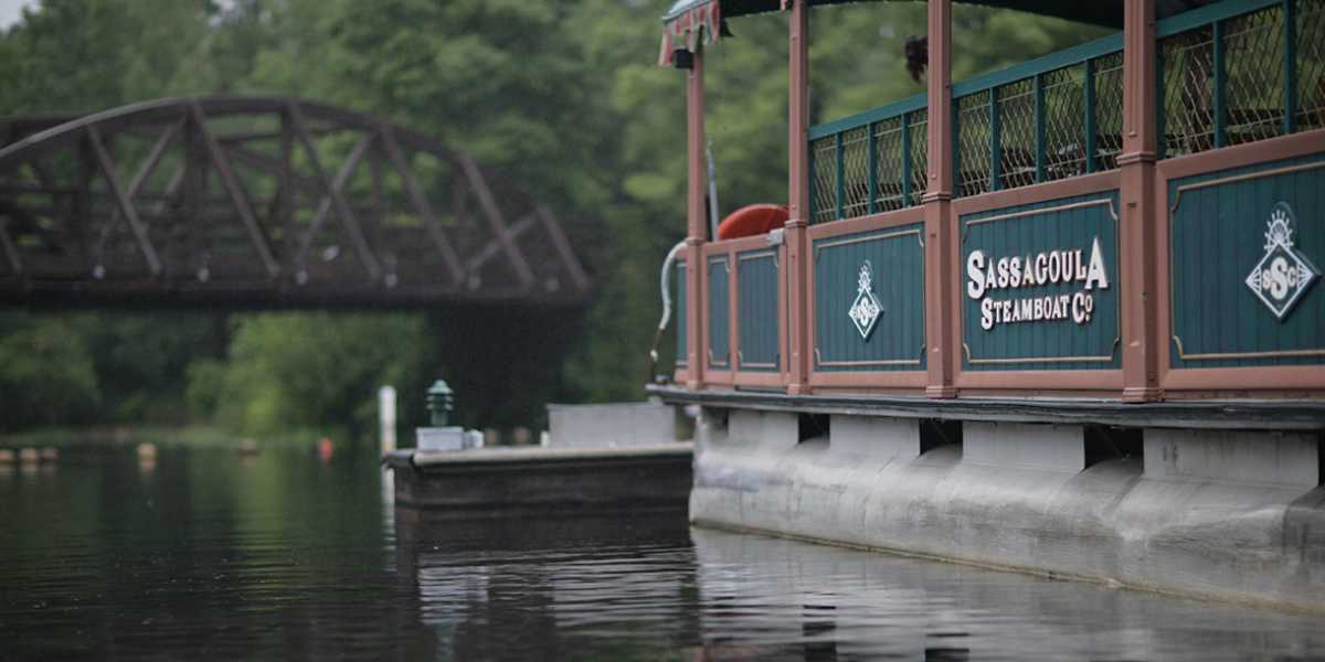 A Disney boat with green and brown colors, displaying the name "Sassagoula Steamboat Co.," is moored on a tranquil river with a trestle bridge partially visible in the background and surrounded by lush green trees, providing charming transportation through scenic landscapes.