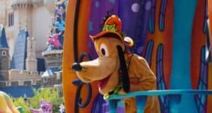 A costumed character resembling Pluto from Disney, dressed in a colorful Halloween-themed outfit with a bright hat, leans over a railing. The backdrop features vibrant decorations including pumpkins and a castle in the background, capturing the magical atmosphere of Tokyo Disney Halloween.