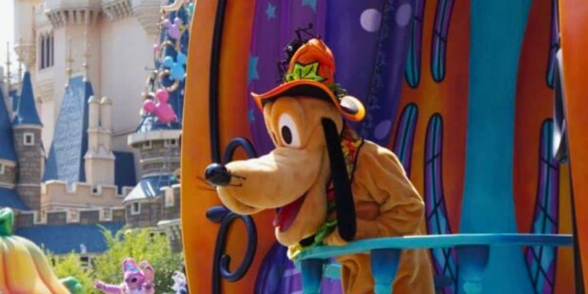 A costumed character resembling Pluto from Disney, dressed in a colorful Halloween-themed outfit with a bright hat, leans over a railing. The backdrop features vibrant decorations including pumpkins and a castle in the background, capturing the magical atmosphere of Tokyo Disney Halloween.