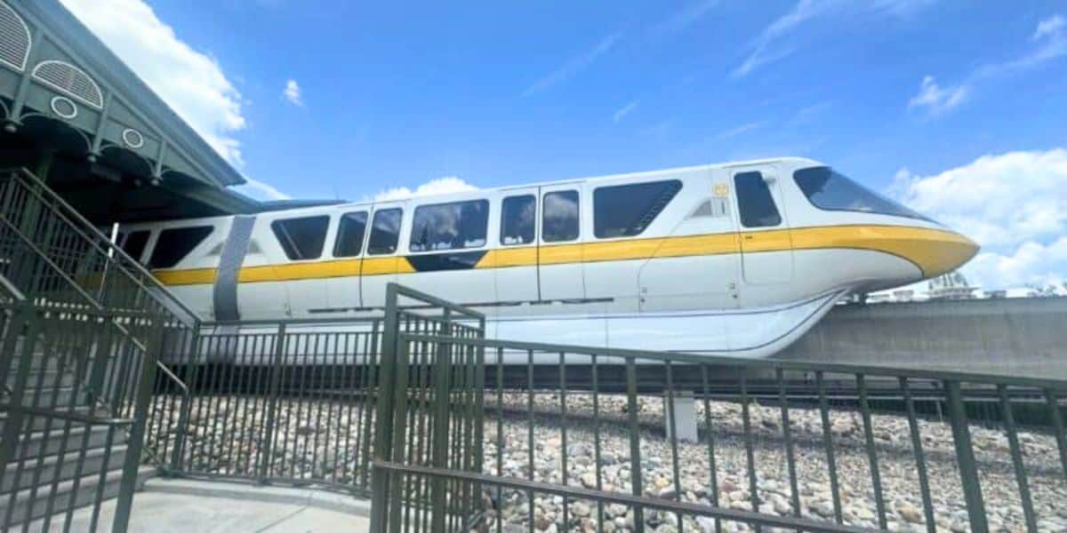 A Disney monorail train in yellow and white colors passing by on an elevated track at a Disney theme park, with a partly cloudy blue sky in the background and a metal fence in the foreground.