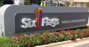 A large sign with colorful flags and bold white text reading "Six Flags World Headquarters" stands amid manicured landscaping with red flowers and green bushes, signifying the new policies on guest access. A building and a parked vehicle are visible in the background.