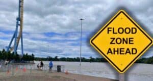 A large yellow sign reads "FLOOD ZONE AHEAD" in bold black letters. Behind the sign, Valleyfair amusement park appears partially submerged by floodwaters under a cloudy sky. A few people can be seen walking on the elevated ground near the water's edge at Valleyfair theme park.