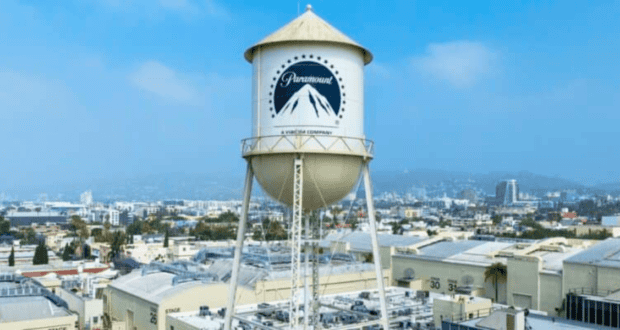 A prominent water tower with the Paramount Pictures logo stands tall against a backdrop of an urban landscape under a bright sky. The surrounding area features various industrial buildings and city structures in the distance, reminiscent of iconic scenes from classic Disney films.