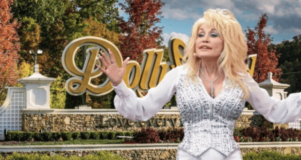 A blonde woman in a sparkly white outfit stands with her arms outstretched in front of the Dollywood sign. The background features trees with autumn foliage and a stone wall below the sign that reads "Dollywood" with the tagline "Love every moment." The scene embodies the spirit of Dolly Parton.