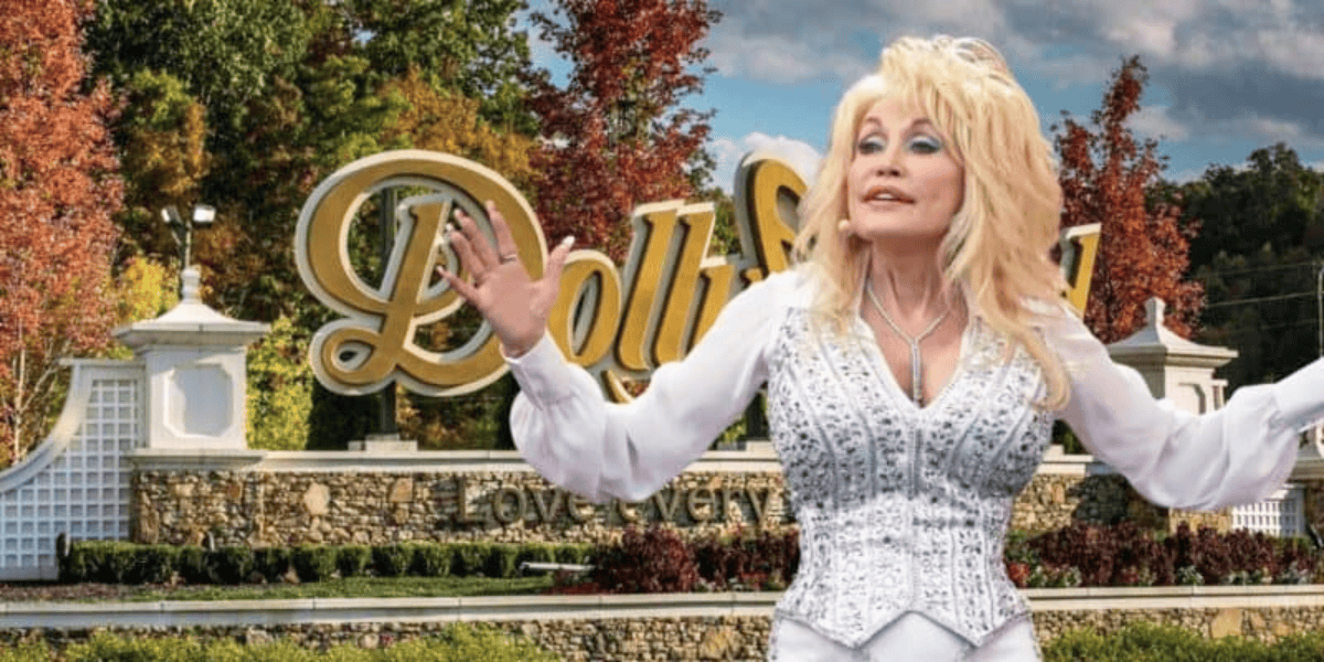 A blonde woman in a sparkly white outfit stands with her arms outstretched in front of the Dollywood sign. The background features trees with autumn foliage and a stone wall below the sign that reads "Dollywood" with the tagline "Love every moment." The scene embodies the spirit of Dolly Parton.