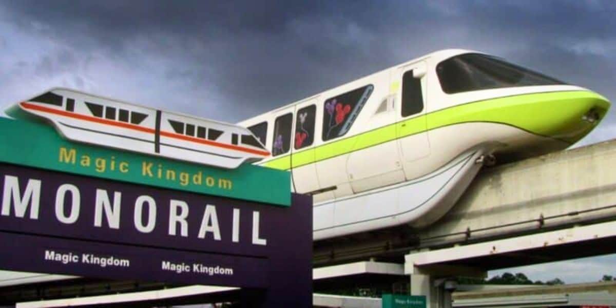 A green and white monorail glides along an elevated track under a cloudy sky. In the foreground, a purple sign with the words "Magic Kingdom Monorail" and an illustration of the monorail indicates the entrance to the transportation system.