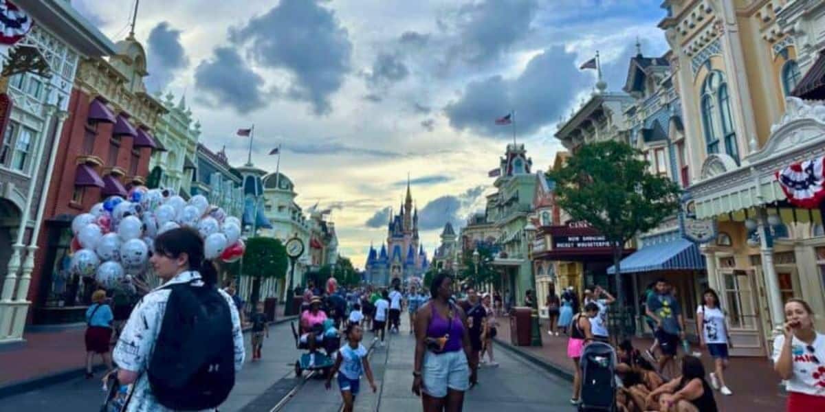 A bustling street scene at a theme park featuring people walking down the main street with the iconic castle in the background. Balloons, decorated shopfronts, and flags add to the festive atmosphere. The sky is partly cloudy, capturing a lively and vibrant moment.