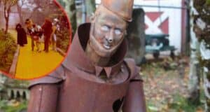 A close-up of a rusted Tin Man statue, with peeling paint and visible wear, set in an outdoor area surrounded by trees. In the top left corner, there is an inset of characters from "The Wizard of Oz" walking on the yellow brick road.