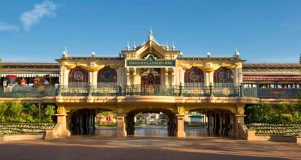 The image shows the main entrance of Disneyland Paris under a bright, clear sky. The building features ornate architecture with a sign reading "Disneyland Paris" and is decorated with gold-painted accents and floral arrangements. In the background is the Disneyland Railroad.