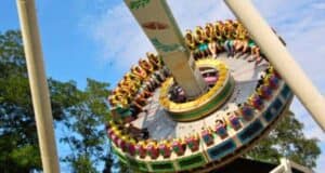People are sitting in a circular amusement ride at a theme park, secured by harnesses, as it moves. The ride is tilted at an angle against the blue sky and trees in the background. Bright colors decorate the ride, and the participants seem engaged in the thrilling experience.