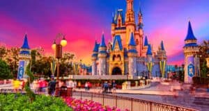 A vibrant scene of Cinderella Castle at sunset, with a stunning sky of pink, purple, and orange hues. Visitors are walking around the castle, which is adorned with blue roofs and golden accents. Bright flowers and decorative medieval banners enhance the foreground as the park reopens to delighted crowds.