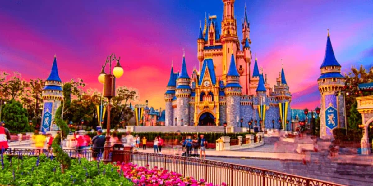 A vibrant scene of Cinderella Castle at sunset, with a stunning sky of pink, purple, and orange hues. Visitors are walking around the castle, which is adorned with blue roofs and golden accents. Bright flowers and decorative medieval banners enhance the foreground as the park reopens to delighted crowds.