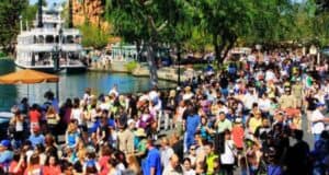A busy outdoor scene at a Disney theme park with a large crowd of people, Disney merchandise stalls, a riverboat in the background, trees, and a clear blue sky.