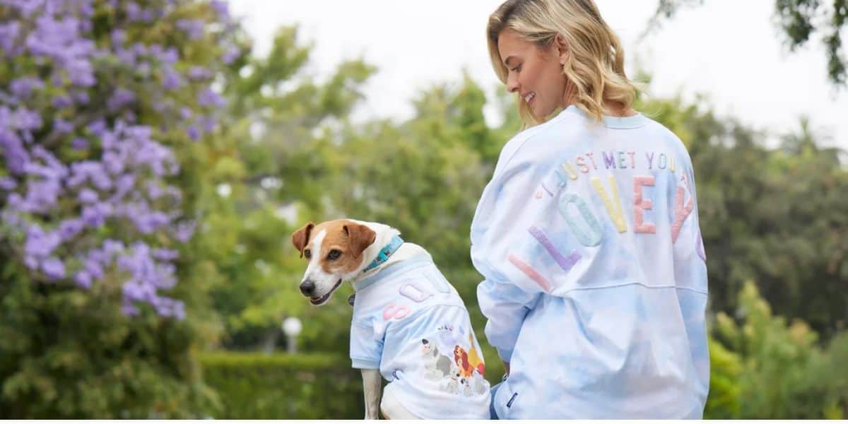 A woman with blonde hair is outdoors, wearing a light blue sweatshirt that says "I just met you, I love you" on the back. She is smiling and looking at her dog, who’s also in a matching light blue shirt. Purple flowers and greenery set the scene, creating a moment straight out of Disney World.