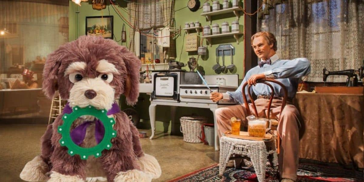 A plush dog toy with a green ring in its mouth sits on the floor of a vintage-style kitchen with various kitchenware and utensils. Nearby, a mannequin of a man in period attire holds a glass of iced tea and sits in a chair near an oven, creating a nostalgic scene reminiscent of Disney World.