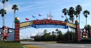 Walt Disney World entrance with a large archway reading "Walt Disney World: Where Dreams Come True." Minnie Mouse is on the left, Mickey Mouse is on the right. Palm trees and a castle-like structure are visible in the background.