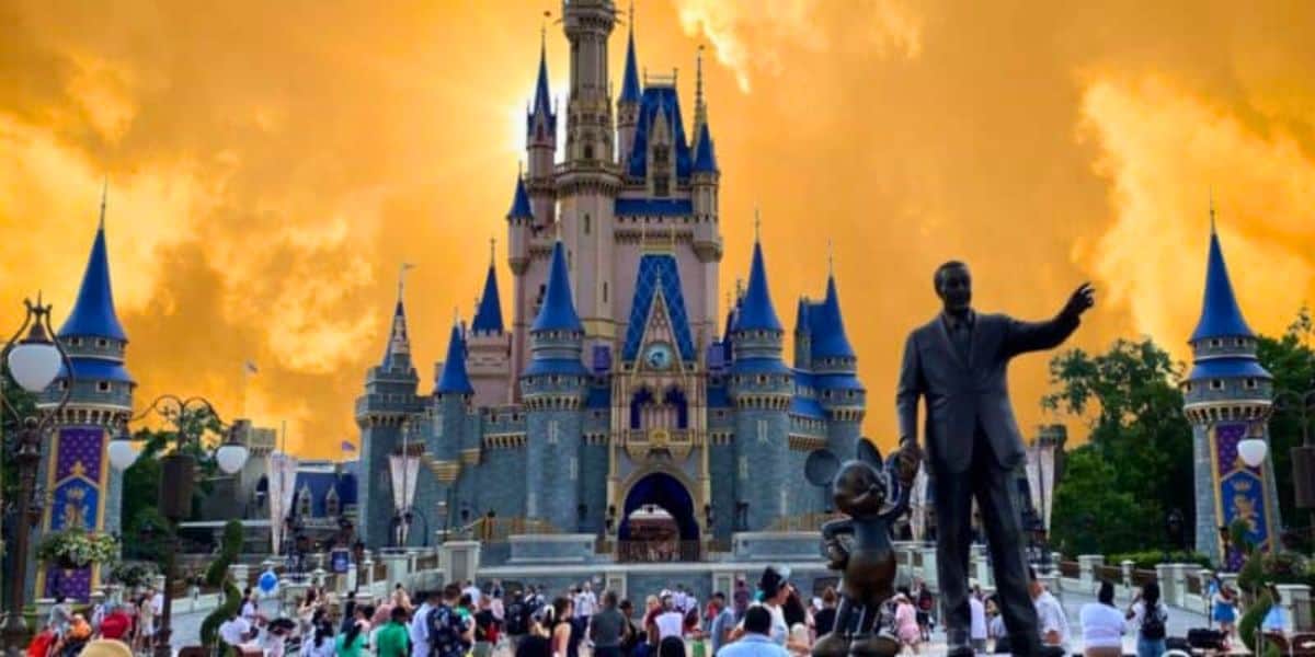 Sunset hues cast a dramatic backdrop over the iconic cinderella castle and the statue of walt disney and mickey mouse at a bustling disney theme park.