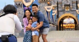 A family of four, consisting of two adults and two children, poses for a photo in front of a castle at a themed amusement park. The woman wears Minnie Mouse ears with a bow, and they all smile happily while hugging each other.