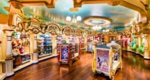 A brightly lit, colorful toy store with wooden floors and ornate architecture. Shelves are lined with a variety of toys, including stuffed animals and action figures. Display stands in the center feature themed Disney merchandise. A painted sky ceiling adds to the whimsical atmosphere.