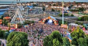 A large outdoor music festival with a massive crowd gathered in front of a stage. An amusement park, particularly popular for its Ferris wheel and roller coaster, looms in the background, surrounded by trees and buildings. The image is taken from an elevated vantage point.