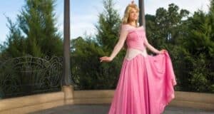 A woman dressed as a princess, wearing a pink ball gown, smiling and posing on a gazebo with green trees in the background.