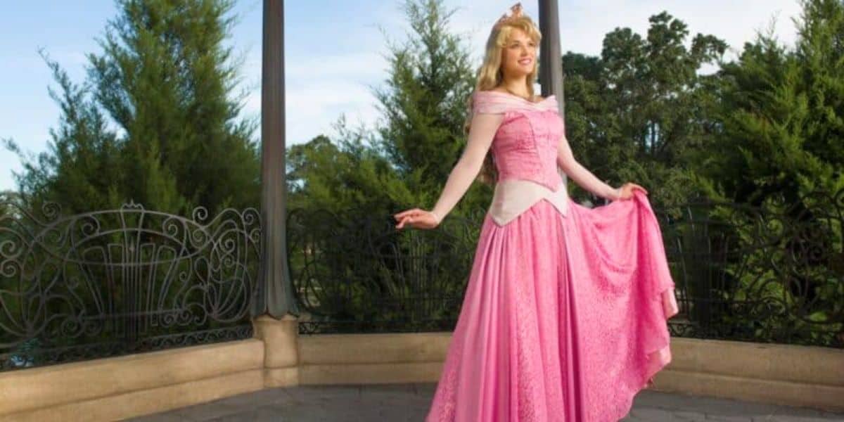 A woman dressed as a princess, wearing a pink ball gown, smiling and posing on a gazebo with green trees in the background.