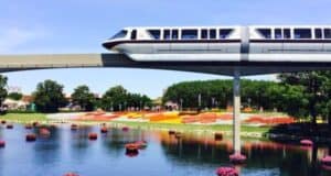 A monorail glides over a scenic park with vibrant flower beds and floating floral decorations on a sunny day.
