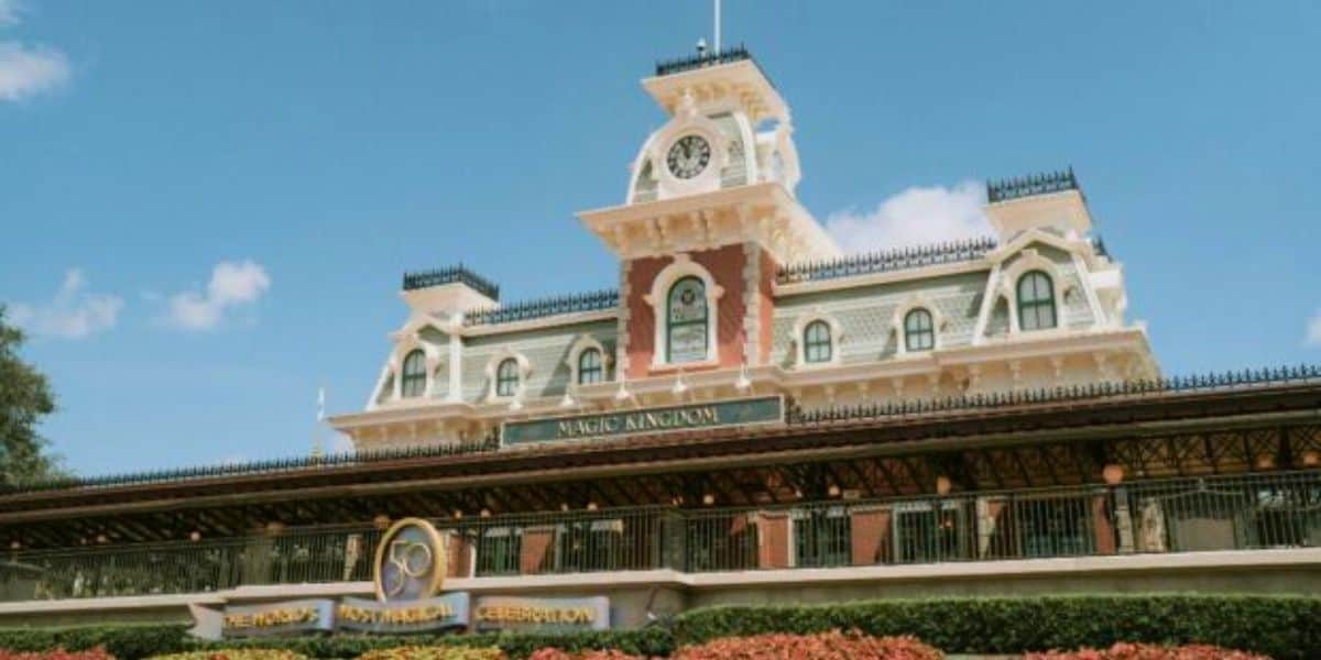 The image captures the entrance to a theme park, reminiscent of Disney magic, with a grand Victorian-style train station and clock tower. Decorative elements stand out against a clear blue sky, while lush landscaping with red flowers adorns the foreground. Nearby, Disney Child Care Services ensures family-friendly fun.
