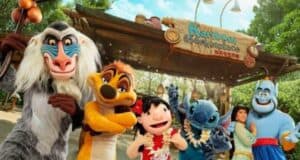 A group of people dressed as Disney characters, including a baboon, a meerkat, Stitch, the Genie, and others, are posing for a photo in front of a sign that reads "Karibuni Marketplace." The background features lush greenery and a wooden structure.