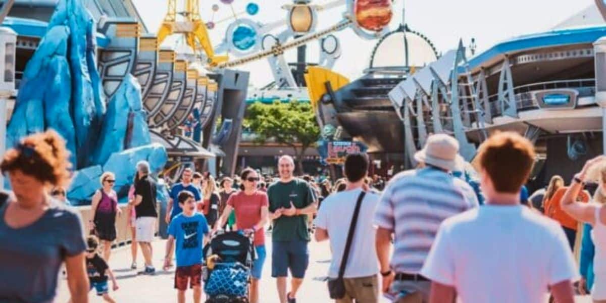 A bustling theme park with many visitors walking along a wide path. Colorful attractions, including futuristic structures and rides, are in the background, and people of all ages, including families, are enjoying their day in bright, sunny weather.