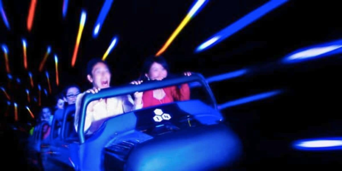A group of people are riding a roller coaster in a dark environment with colorful light streaks creating a sense of motion and excitement. The passengers appear thrilled, with some screaming and others smiling as they hold onto the safety bar, enjoying the iconic Disney experience on Space Mountain.