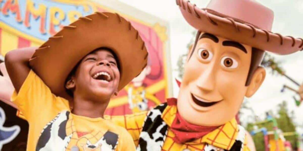 A child wearing a cowboy hat and a yellow shirt laughs joyfully next to a person in a Woody costume from Toy Story. They are at an amusement park with colorful signs in the background, creating a lively and fun atmosphere.