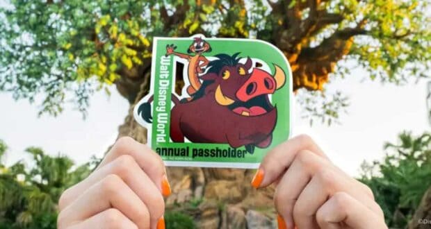 A person with orange-painted nails holds up an annual passholder sticker featuring Timon and Pumbaa from Disney’s The Lion King. The background features lush greenery and the iconic Tree of Life from Disney's Animal Kingdom.