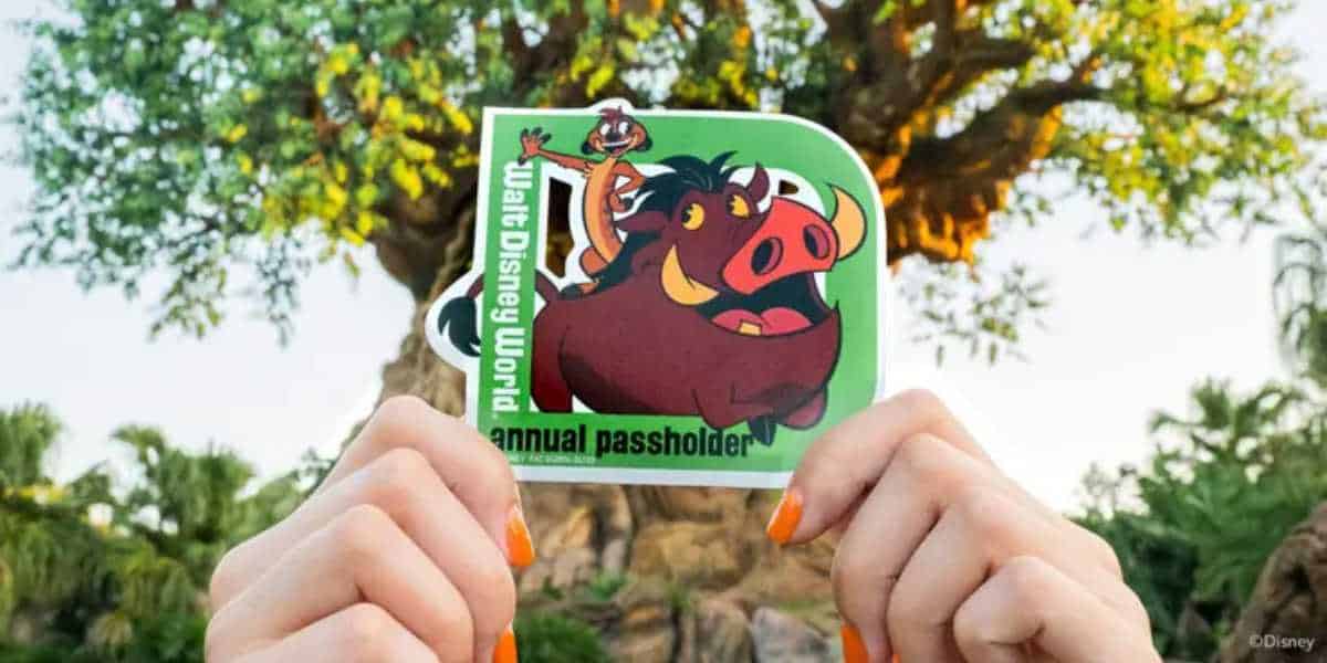 A person with orange-painted nails holds up an annual passholder sticker featuring Timon and Pumbaa from Disney’s The Lion King. The background features lush greenery and the iconic Tree of Life from Disney's Animal Kingdom.