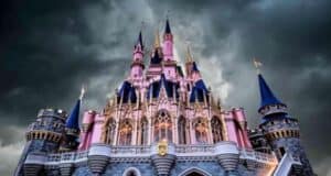 A majestic castle with tall spires and intricate details, illuminated under dramatic dark clouds. The castle is primarily pink with blue rooftops and flags, creating a striking contrast with the stormy sky.