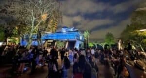A large crowd of people gathers at a theme park at night. The area is illuminated with blue and yellow lights, highlighting a futuristic building in the background. Trees and greenery surround the scene, with a partly cloudy sky overhead.
