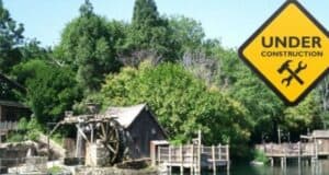 A wooden mill house with a large water wheel stands near a river, surrounded by dense greenery. A yellow diamond-shaped "Under Construction" sign with crossed tools appears in the sky area of the image on the right side, indicating ongoing construction.