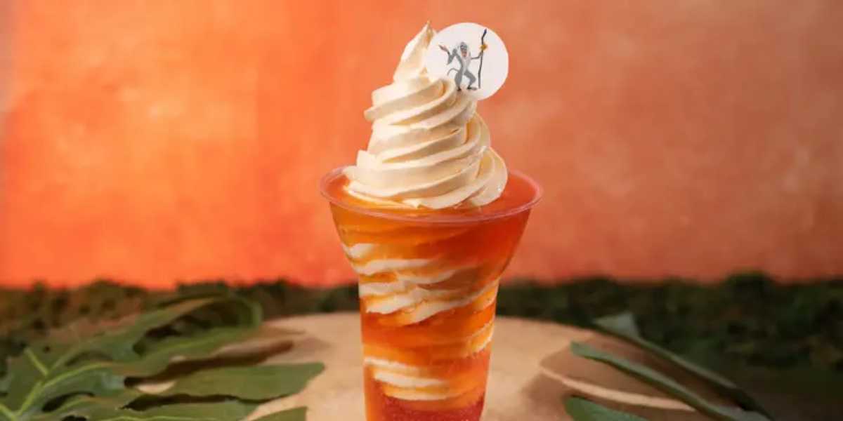 An image of a tall, swirled soft-serve ice cream in a clear cup. The ice cream is a mix of white and orange hues and sits atop a layered red and orange base. The cup is garnished with a small decorative pick featuring a character design. The background is blurred.