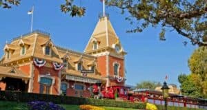 The iconic Disneyland Railroad train station is seen with a classic red train at the platform, adorned with red, white, and blue bunting. The building is surrounded by well-maintained landscaping, flowers, and trees, with blue skies and a flag atop the roof.