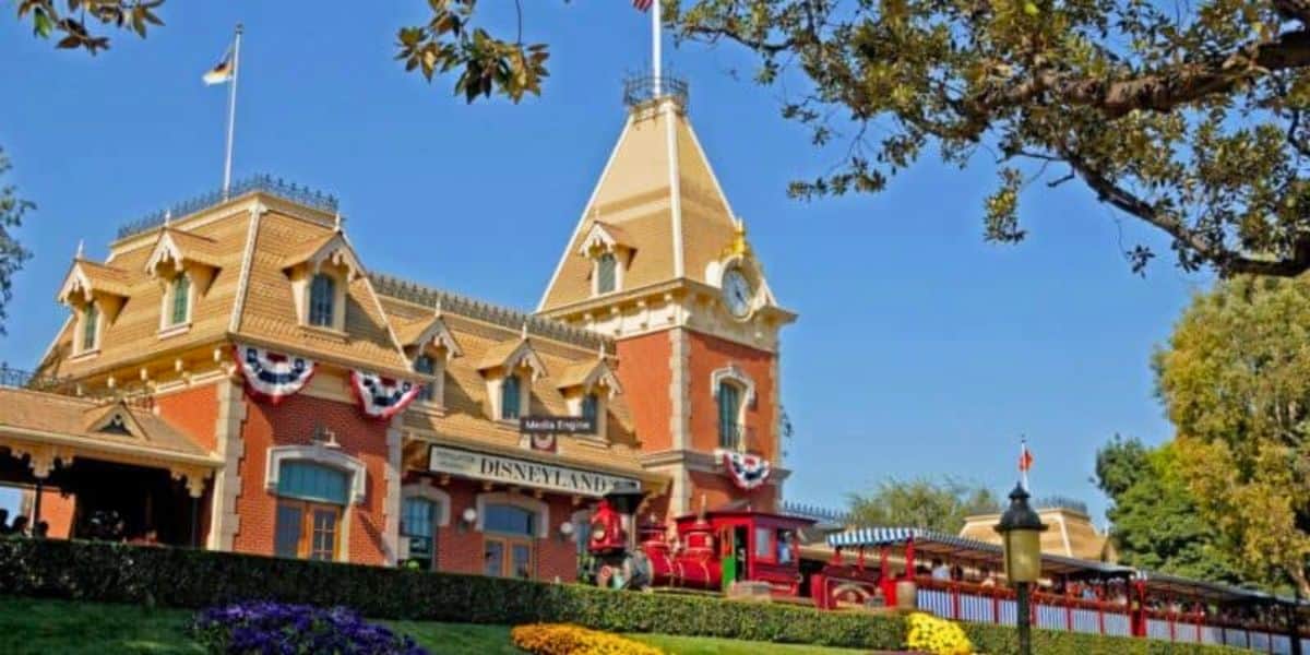 The iconic Disneyland Railroad train station is seen with a classic red train at the platform, adorned with red, white, and blue bunting. The building is surrounded by well-maintained landscaping, flowers, and trees, with blue skies and a flag atop the roof.