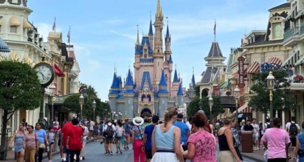 A bustling crowd walks down a wide street lined with shops and trees towards a large, ornate castle with spires and blue roofs, reminiscent of Disney World Vacations. The sky is clear, and people are enjoying their day in what appears to be a theme park.