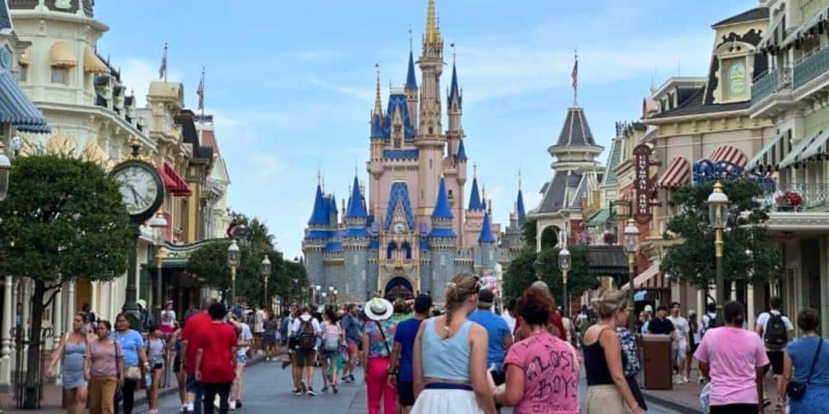 A bustling crowd walks down a wide street lined with shops and trees towards a large, ornate castle with spires and blue roofs, reminiscent of Disney World Vacations. The sky is clear, and people are enjoying their day in what appears to be a theme park.