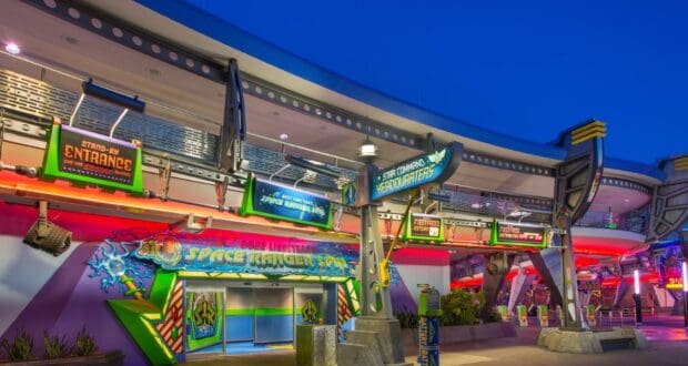 The image captures the entrance to Buzz Lightyear's Space Ranger Spin at a theme park, likely in the early evening. Illuminated by vibrant, colorful signs and lights, the area radiates a futuristic ambiance with space-themed decorations.