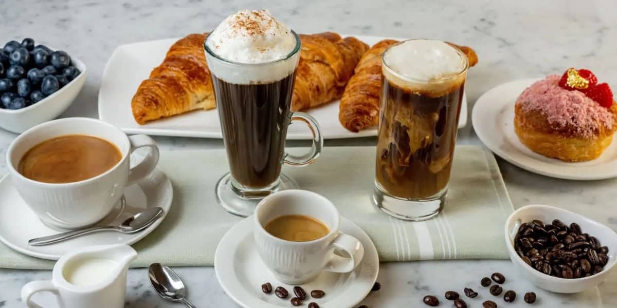 A breakfast spread on a marble surface featuring croissants, a chocolate-filled pastry with a red topping, a bowl of blueberries, and an array of coffees including a small espresso, cappuccino, iced coffee, and black coffee. Coffee beans are scattered around—a feast after tackling Walt Disney World headaches.