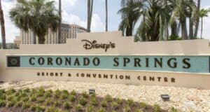 Sign for Disney's Coronado Springs Resort & Convention Center, surrounded by palm trees and greenery. The building in the background complements the sign, which features a light-colored finish and teal accents—a welcoming sight for guests of Walt Disney World.