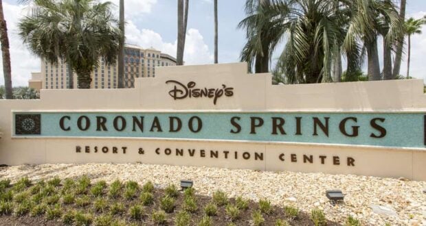 Sign for Disney's Coronado Springs Resort & Convention Center, surrounded by palm trees and greenery. The building in the background complements the sign, which features a light-colored finish and teal accents—a welcoming sight for guests of Walt Disney World.