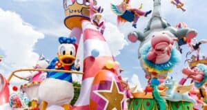 A colorful Disney World parade float features a cartoon duck in a blue sailor outfit with a waving hand. Adorned with whimsical decorations like stars, candy stripes, an elephant in circus attire, and flying fish, this parade float dazzles against the bright blue sky with scattered clouds. Parade viewing tip: arrive early for the best spots!