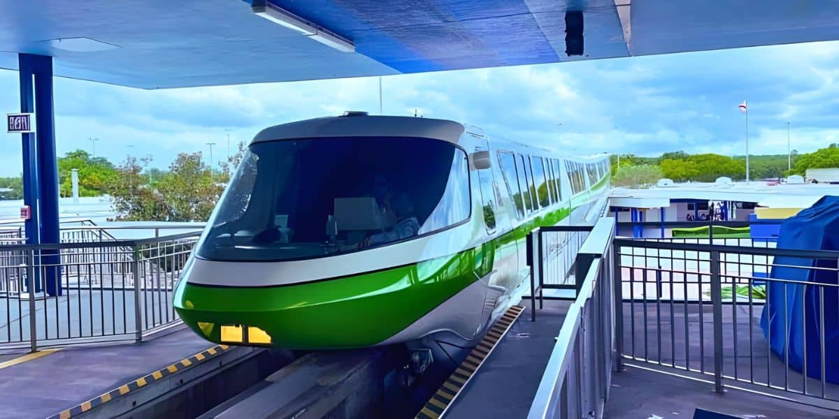 A sleek green and white monorail train arrives at an elevated platform on a sunny day, reminiscent of the Disney Transportation and Ticket Center. The train's streamlined design is visible, with trees and structures in the background. The platform is equipped with railings and safety measures.