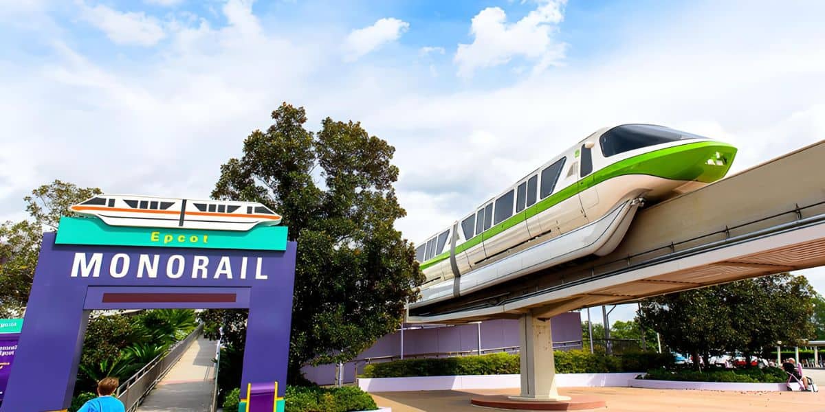 A monorail with green accents glides along an elevated track at Epcot, part of Walt Disney World Resort. The train is passing above a purple sign reading "Epcot MONORAIL" adorned with a monorail graphic. Trees and a cloudy sky complete the scene—the trick to seamless travel? Disney Transportation and Ticket Center.