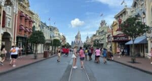 A bustling street scene at Disney World, with a variety of people walking and taking photos. Shops and buildings with decorative facades line both sides. A castle is visible in the distance under a blue sky with scattered clouds.